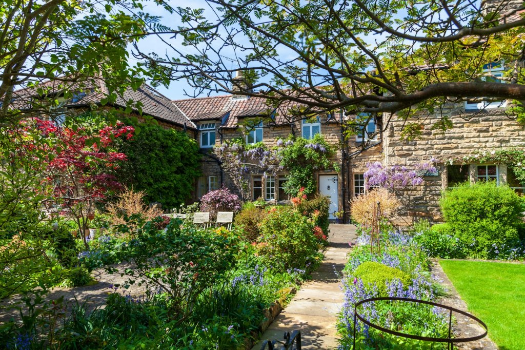 A lush English cottage garden with flower borders and a stone path in front of a house