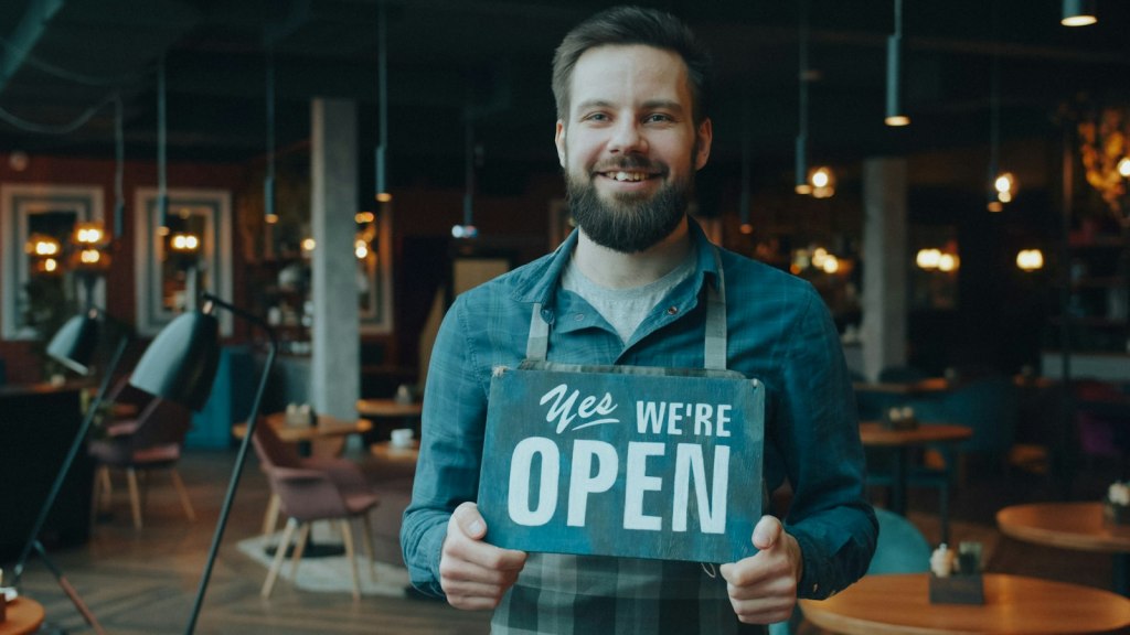 A smiling new business owner holding a Yes we're open sign in his cafe